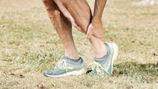 pexels-photo-7298422-7298422 Man holding his calf in pain, wearing teal sneakers on green grass.